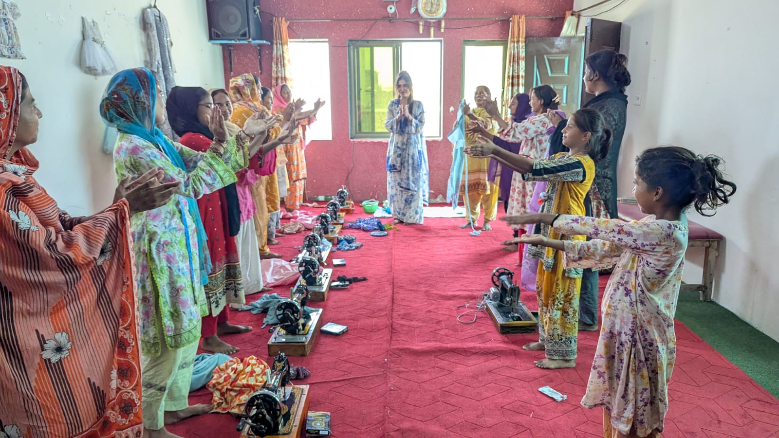 Women and girls in a circle raising their hands in thanks, sewing machines lined up on the floor beside them